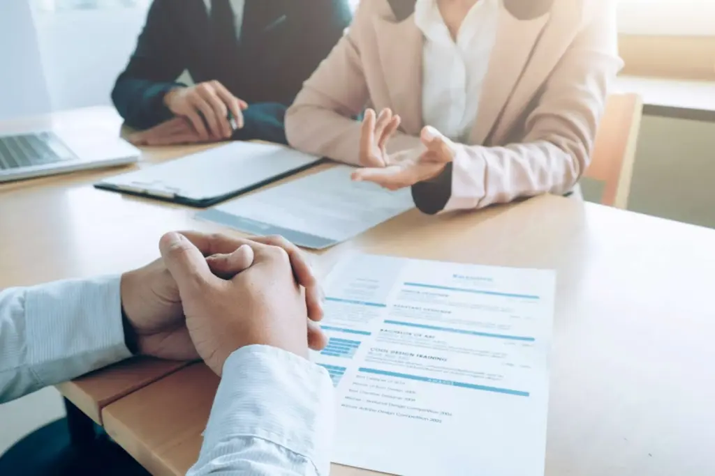 Currículum sobre una mesa de oficina en el contexto de una entrevista de un proceso de selección para cubrir una vacante de empresa. Se ve a dos reclutadores y enfrente las manos del entrevistado sobre su CV.