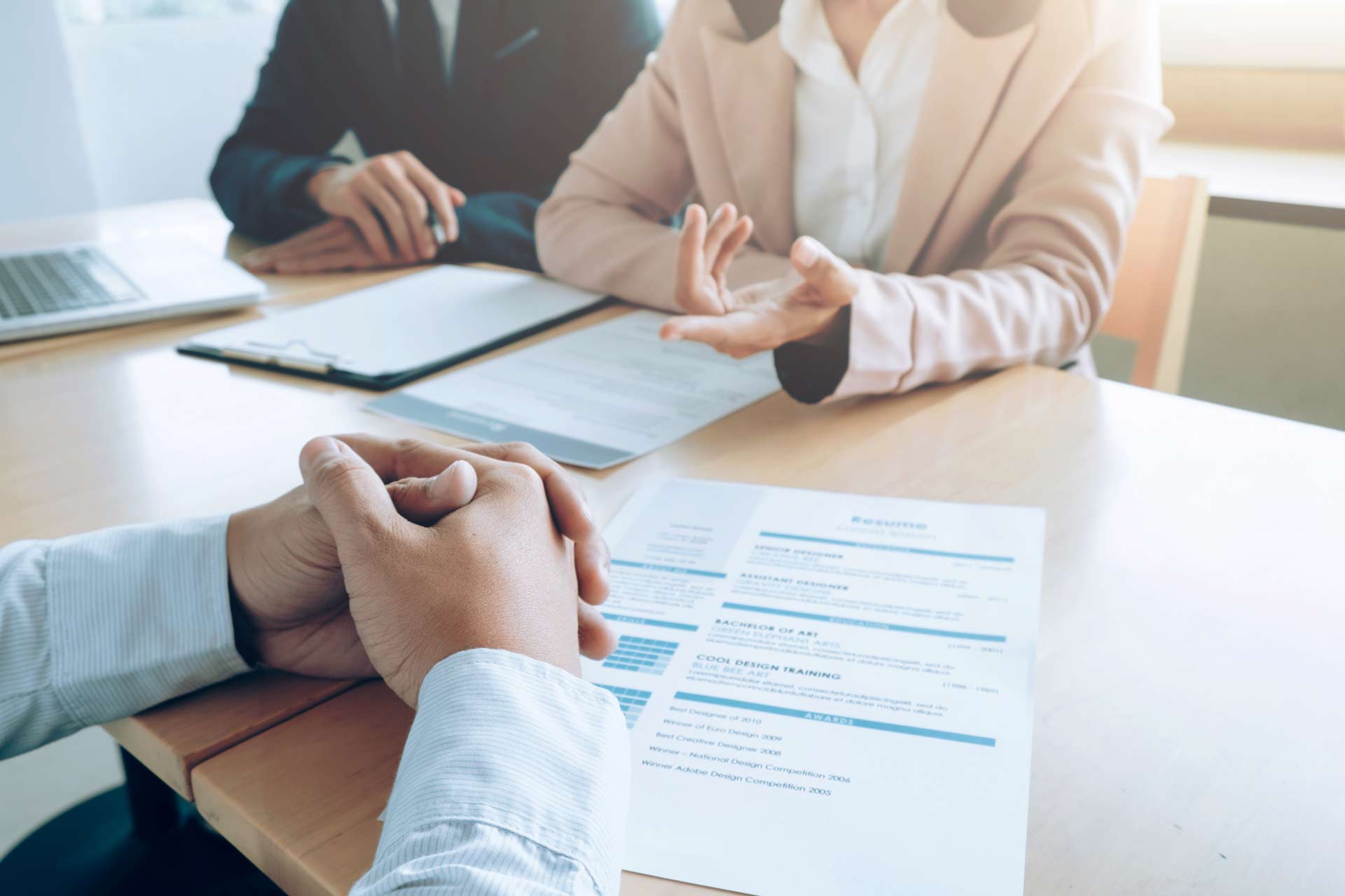 Currículum sobre una mesa de oficina en el contexto de una entrevista de un proceso de selección para cubrir una vacante de empresa. Se ve a dos reclutadores y enfrente las manos del entrevistado sobre su CV.